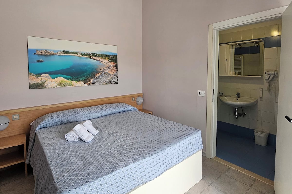 A hotel room with a bed, a painting of a coastal landscape, a bathroom with a sink, and a trash bin.