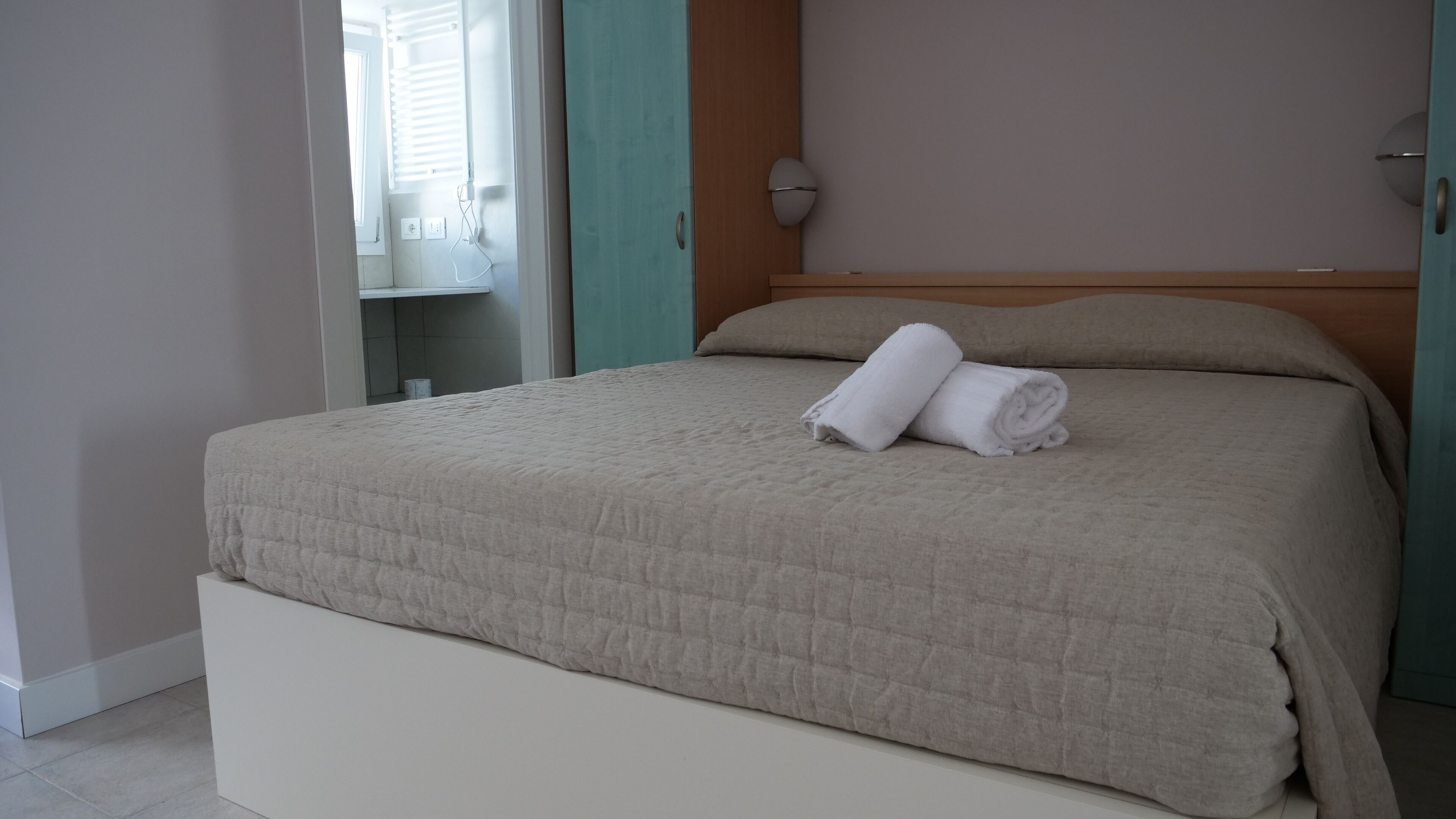 Un lit bien fait, avec une couverture beige et des serviettes blanches pliées, une tête de lit en bois et une petite table de chevet avec une lampe.