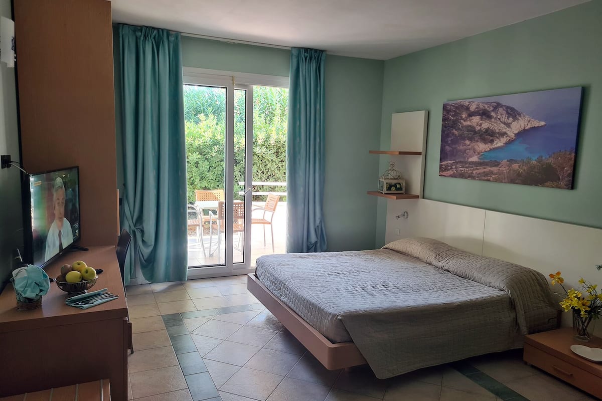 A bedroom with a bed, a desk with a computer, a TV, and a balcony with curtains.