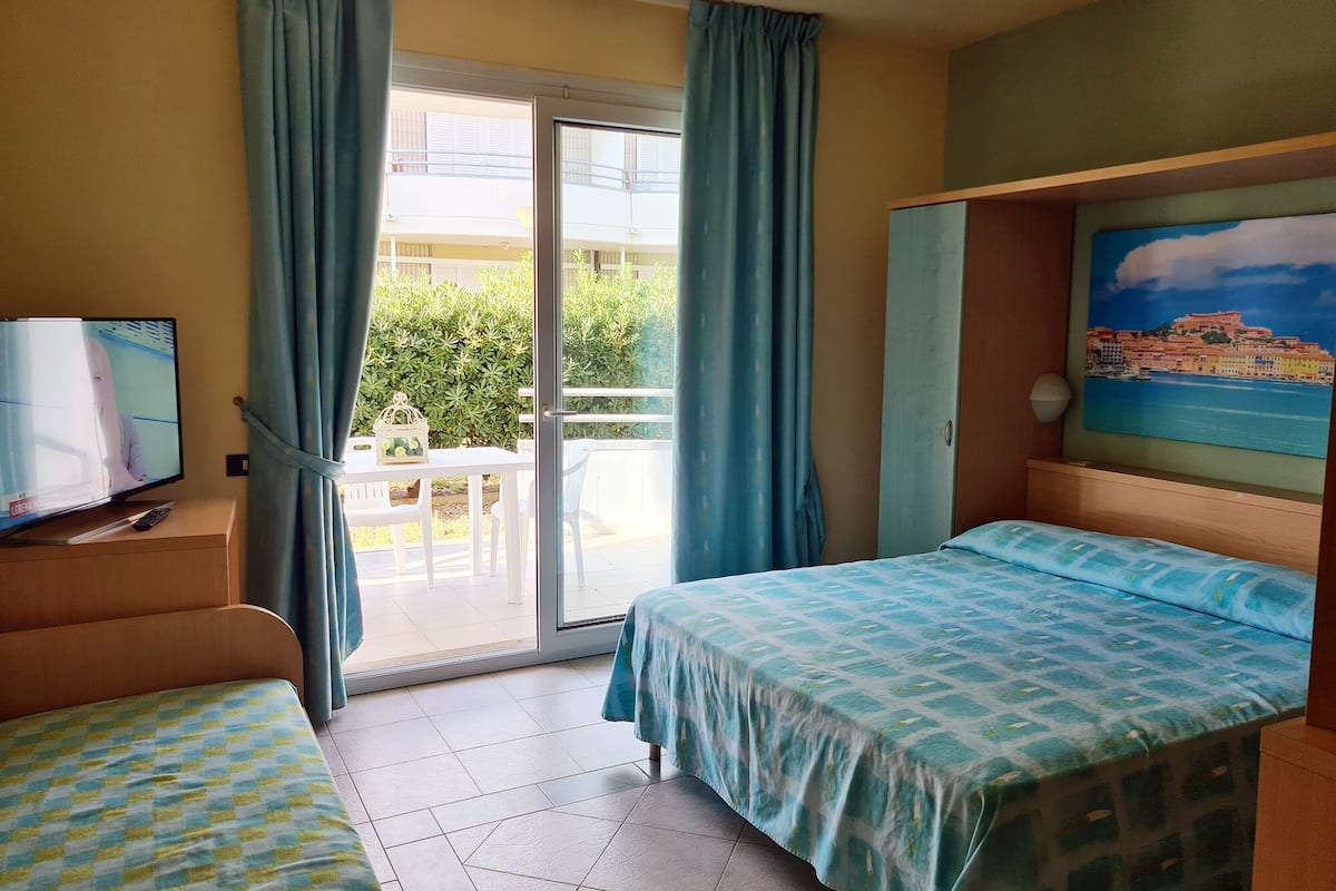 A hotel room with a balcony, a bed with a patterned bedspread, a television on a wooden stand, and a painting of a coastal scene.