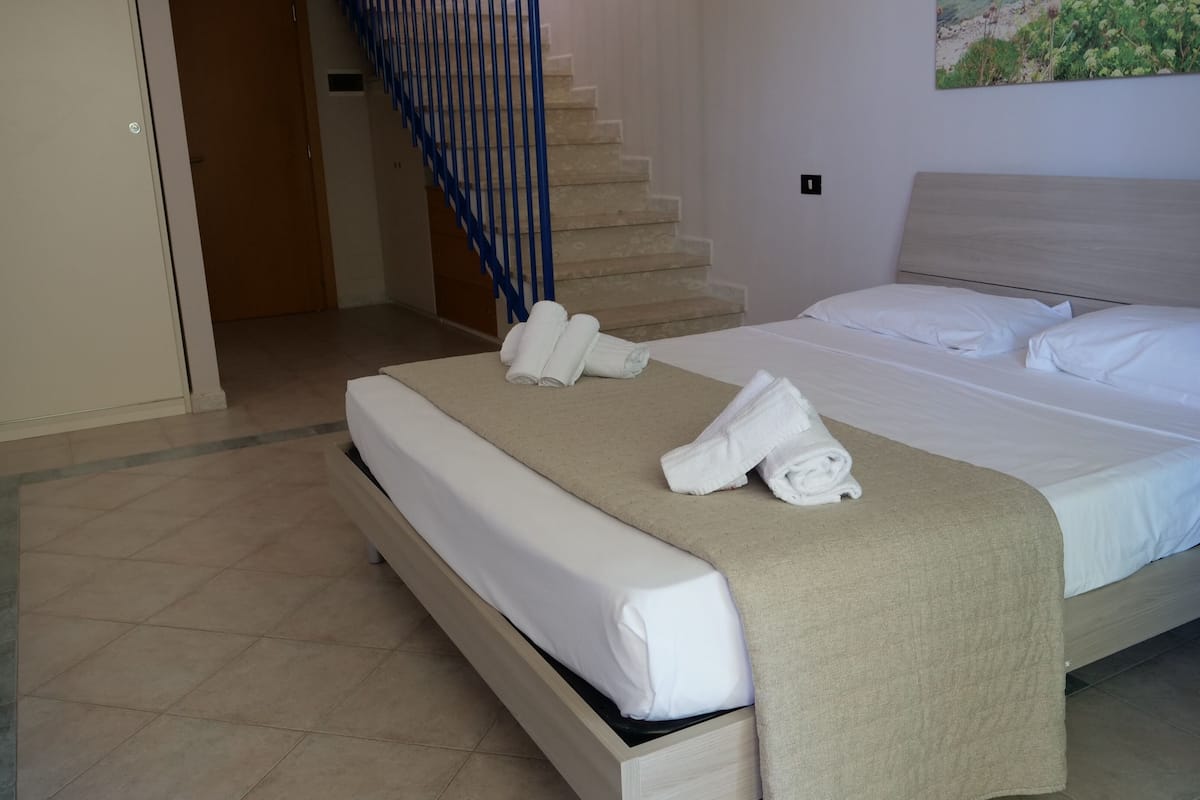 A neatly made bed with white linens and towels, a wooden headboard, and a painting of greenery on the wall.