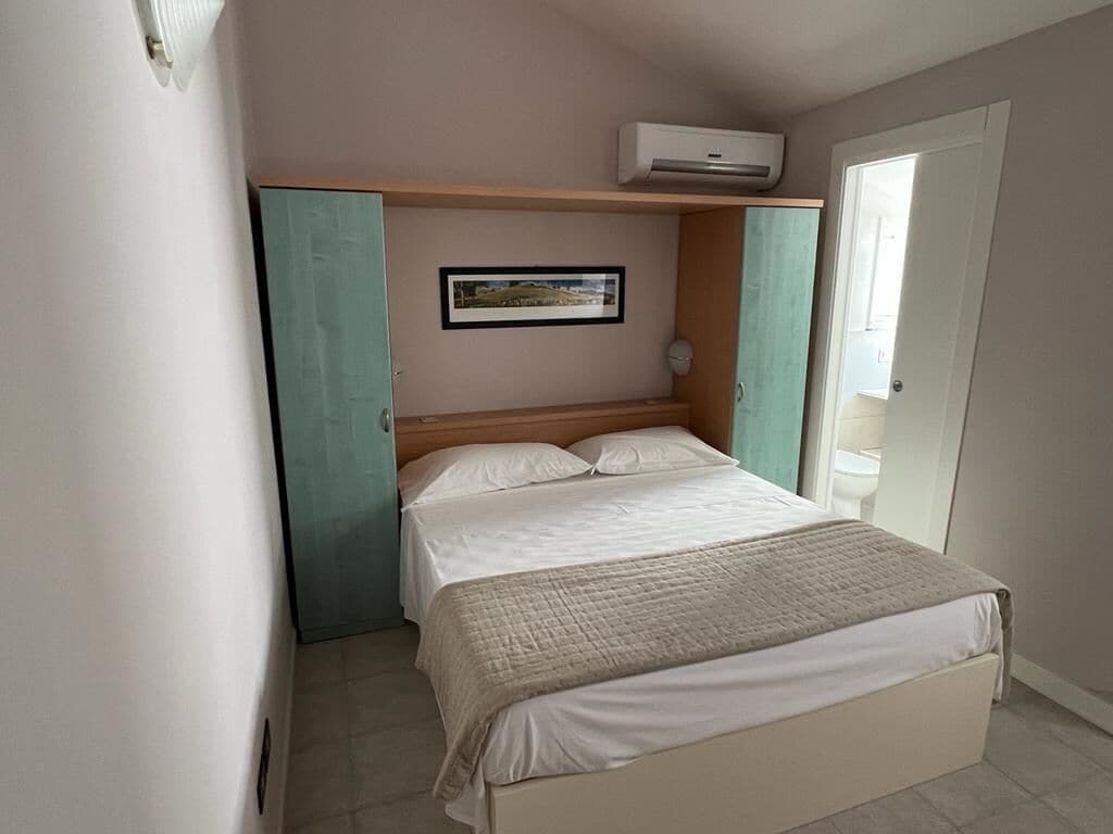 A bedroom with a bed, a wall-mounted air conditioner, a framed picture, and a door.