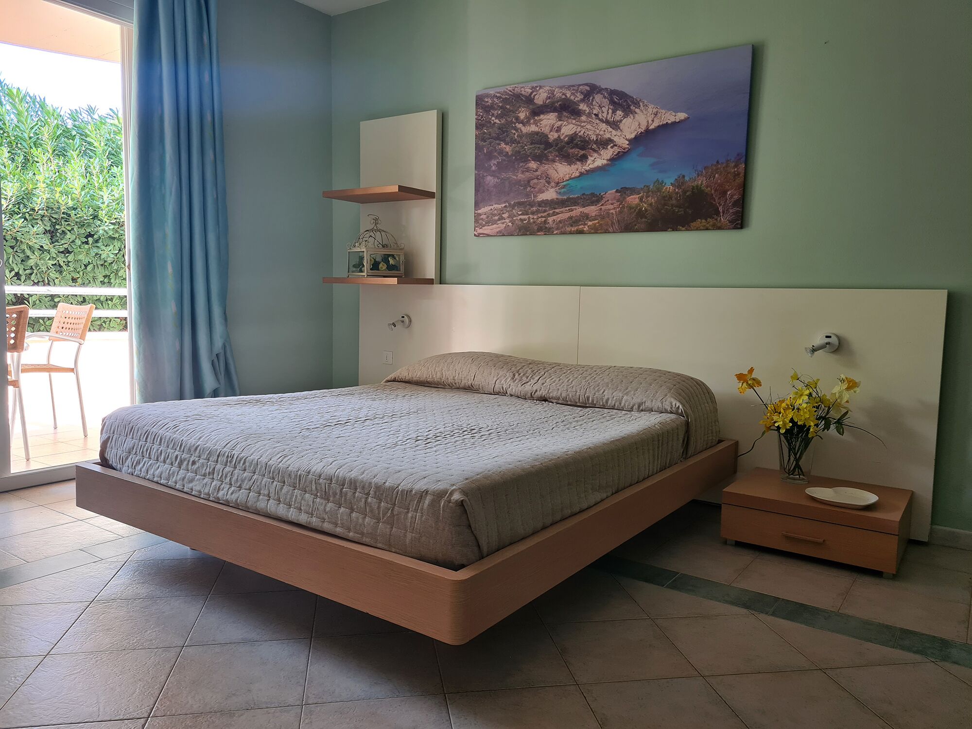 Camera da letto con un letto, un comodino, una mensola a parete, un quadro e vista sul verde.