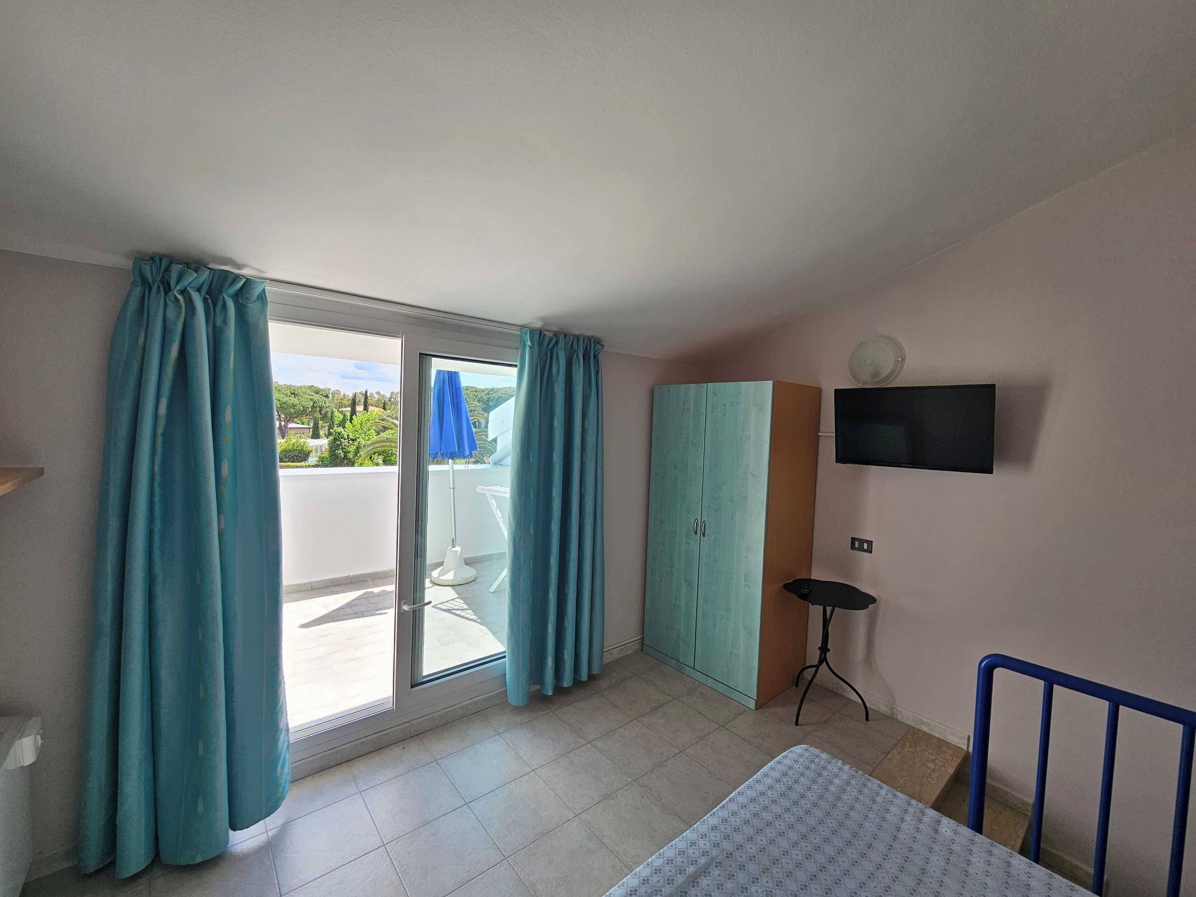 A room with a bed, a TV mounted on the wall, a wardrobe, and a balcony with a blue umbrella.