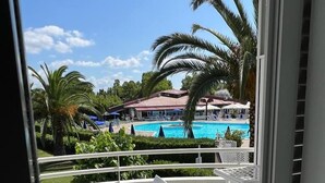 A balcony with a white chair and table, overlooking a swimming pool with lounge chairs and umbrellas, surrounded by palm trees and a building with a red roof. - Hotel La Buca Del Gatto (Cecina)