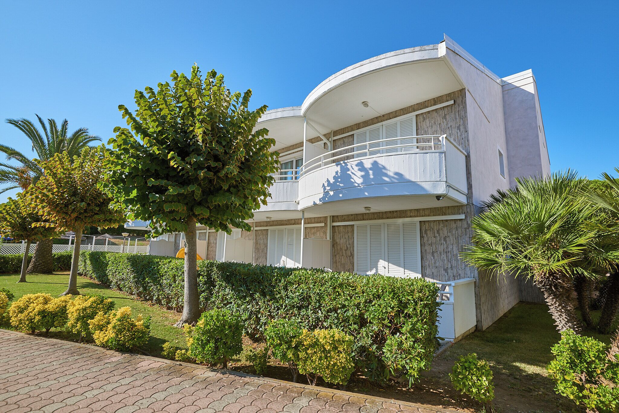 A modern two-story building with balconies, surrounded by well-maintained greenery and a paved walkway.