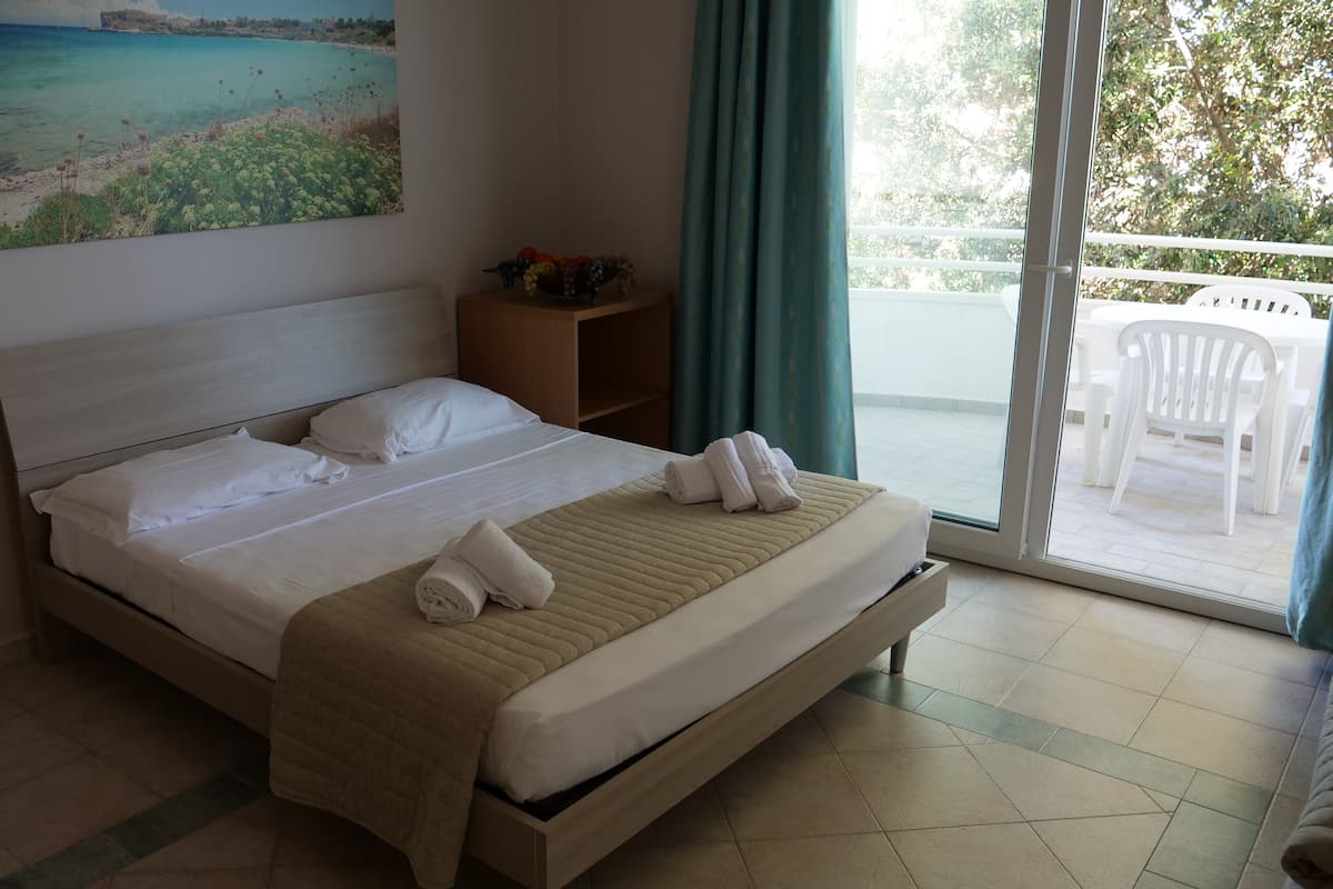 A hotel room with a bed, a painting of a beach, a wooden nightstand, and a view of the outside balcony.