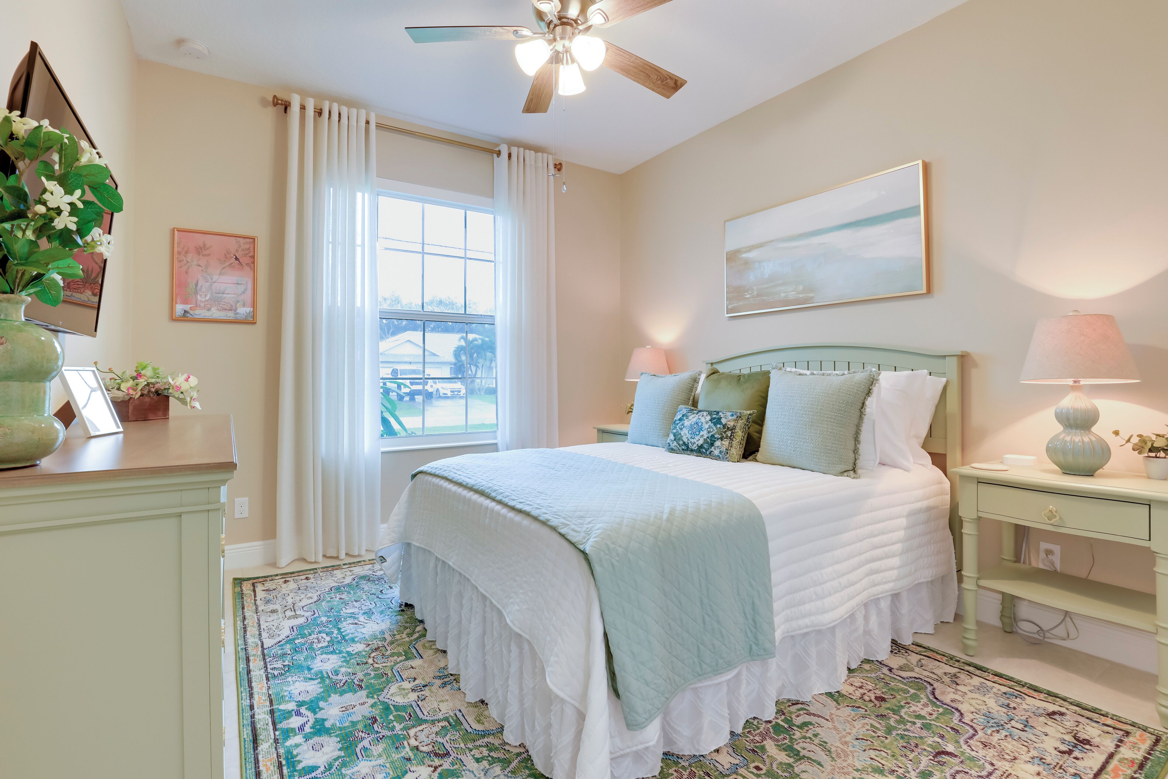 A bedroom with a bed, a nightstand, a lamp, a ceiling fan, a window with curtains, and a rug.