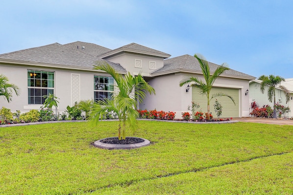 A residential house with a well-maintained lawn, palm trees, and a variety of colorful flowers. - Stylish Port St Lucie Home w/ Hot Tub + Game Room! (Port Saint Lucie)