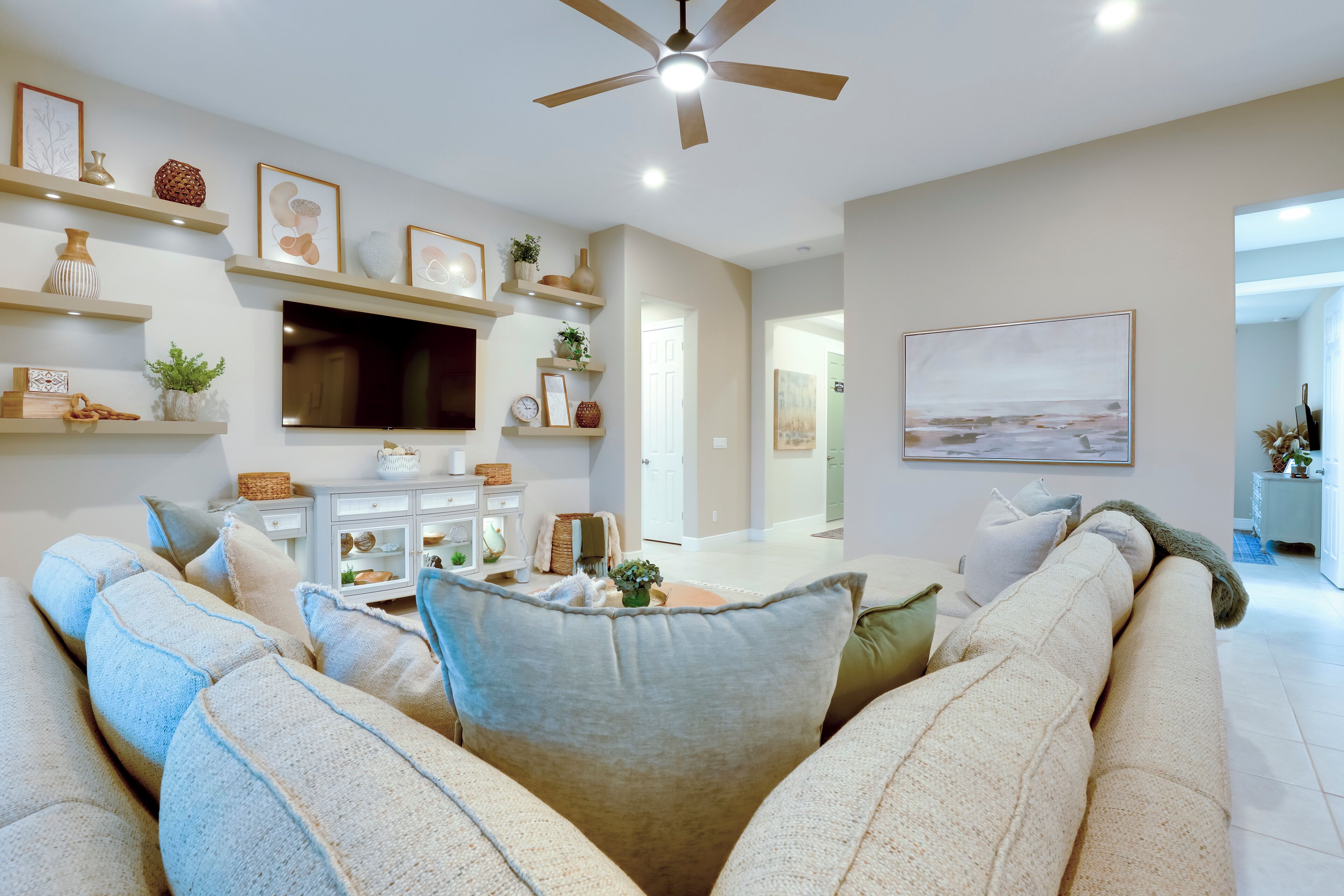A living room with a large sofa, a flat-screen TV, shelves with decorative items, and a ceiling fan.
