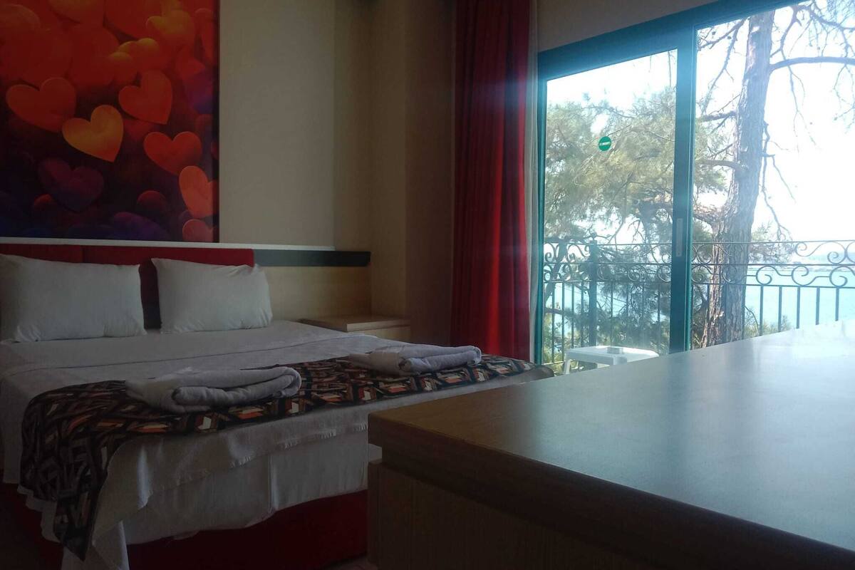 A hotel room with a bed, a large window, and a balcony with a view of trees and a body of water.