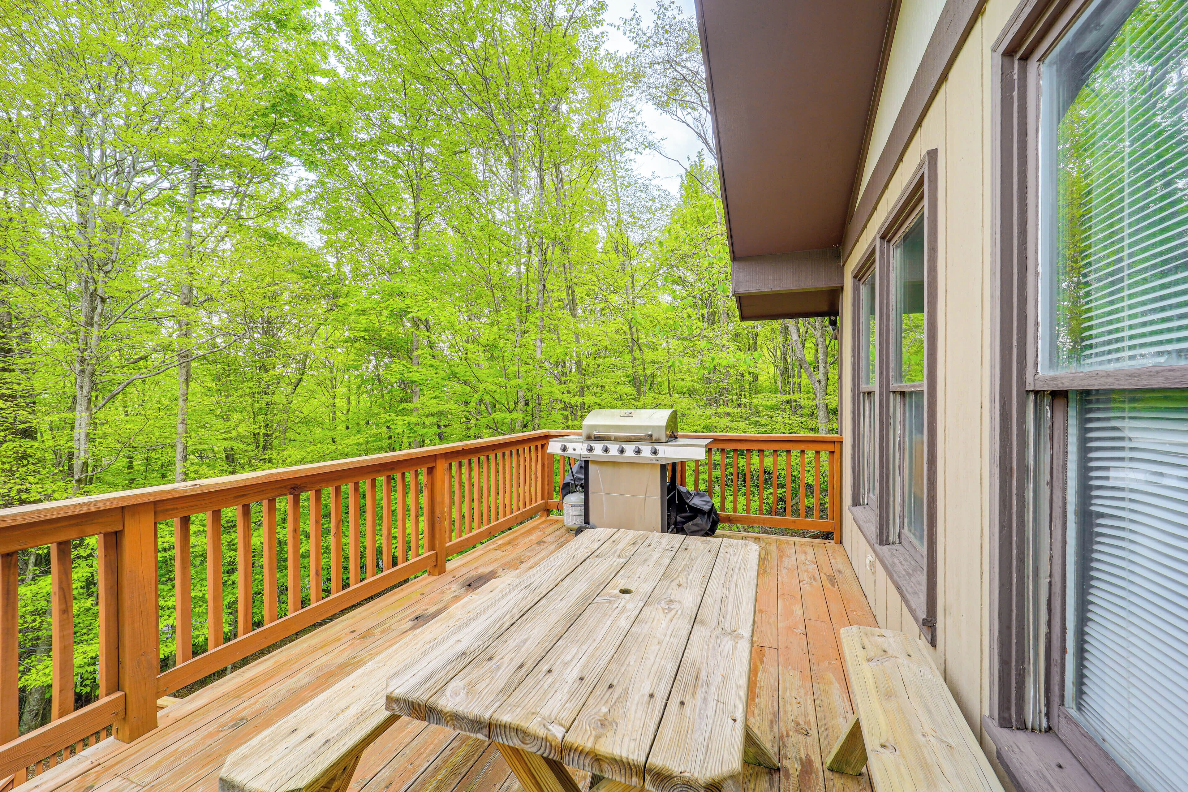 A wooden deck with a grill and table, surrounded by a railing, overlooking a forested area.