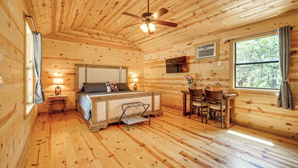 A wooden cabin bedroom with a bed, a desk, a chair, a ceiling fan, and a window. - Cozy Cabin Hill Country Escape w/ Views (Johnson City)