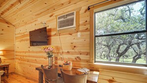 A wooden cabin interior with a dining area, a window with a view of trees, and a wall-mounted air vent. - Cozy Cabin Hill Country Escape w/ Views (Johnson City)