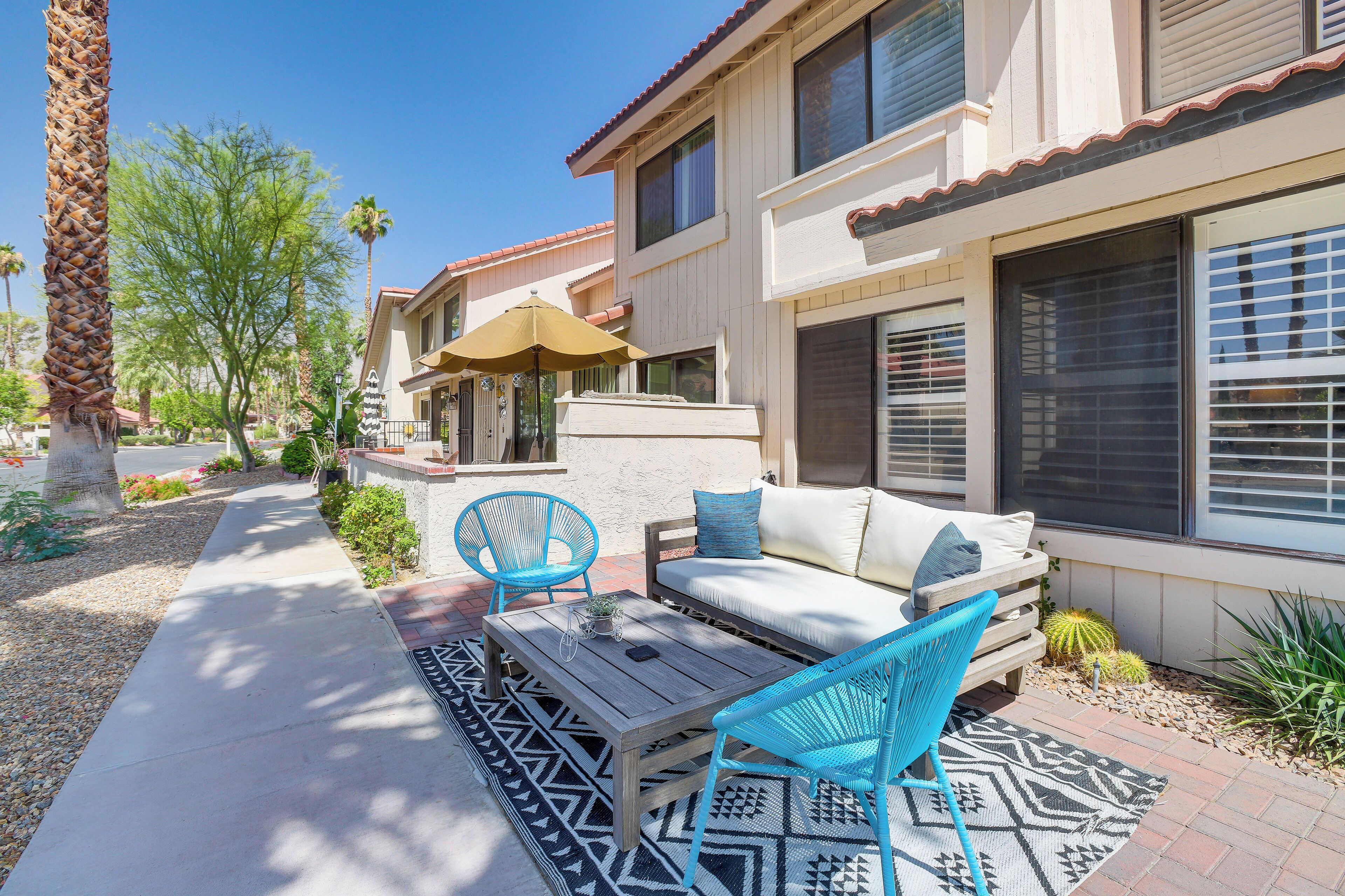 A patio with a wooden table, two blue chairs, a white sofa, and a patterned rug.