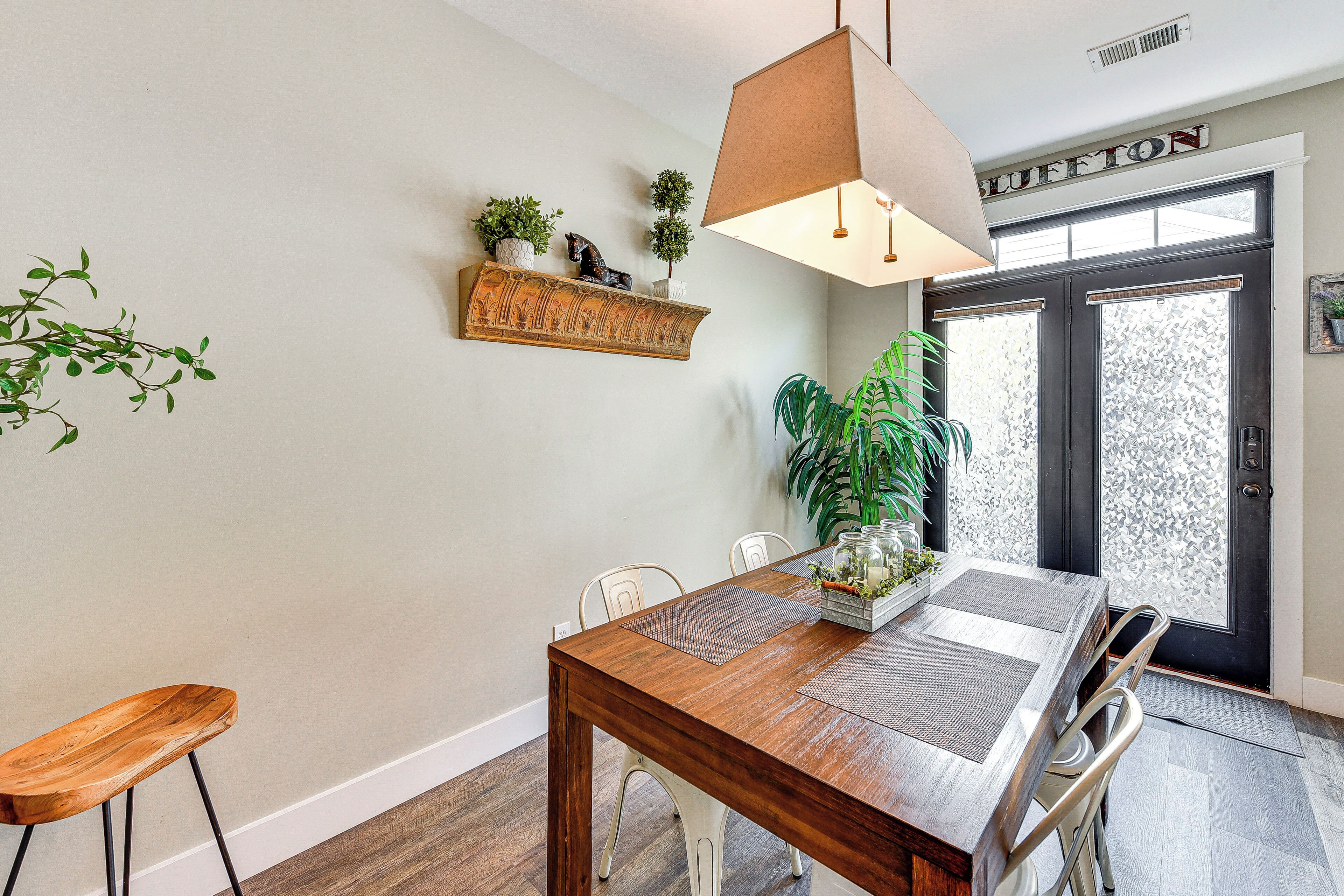 A dining area with a wooden table, chairs, a hanging light fixture, a decorative shelf with plants, and a door with frosted glass panels.