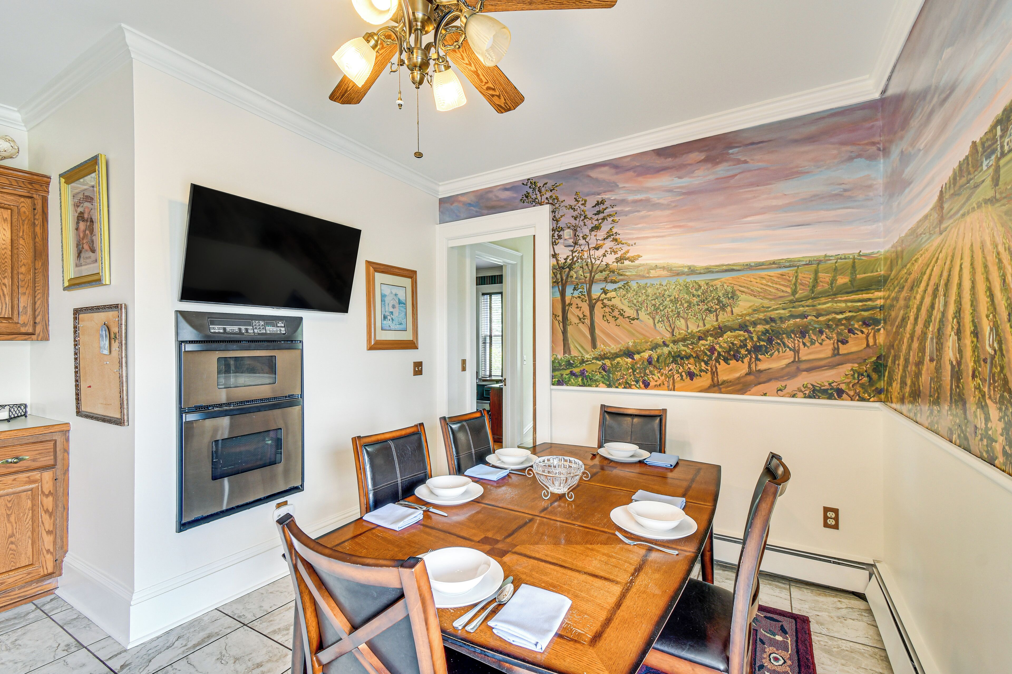 A dining room with a large wooden table set for six, a built-in oven, and a television on the wall.