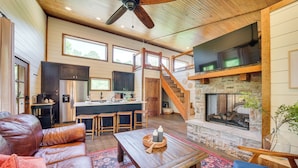 A spacious living area with a fireplace, leather sofa, and a kitchen in the background. - Tree-lined Broken Bow Studio Cabin w/ Hot Tub! (Broken Bow)