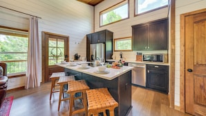 A kitchen with wooden cabinets, a large island with stools, and a view of the outdoors. - Tree-lined Broken Bow Studio Cabin w/ Hot Tub! (Broken Bow)