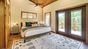 A bedroom with a bed, bedside tables, a ceiling fan, a patterned rug, and a view of the outdoors through a sliding door. - Tree-lined Broken Bow Studio Cabin w/ Hot Tub! (Broken Bow)