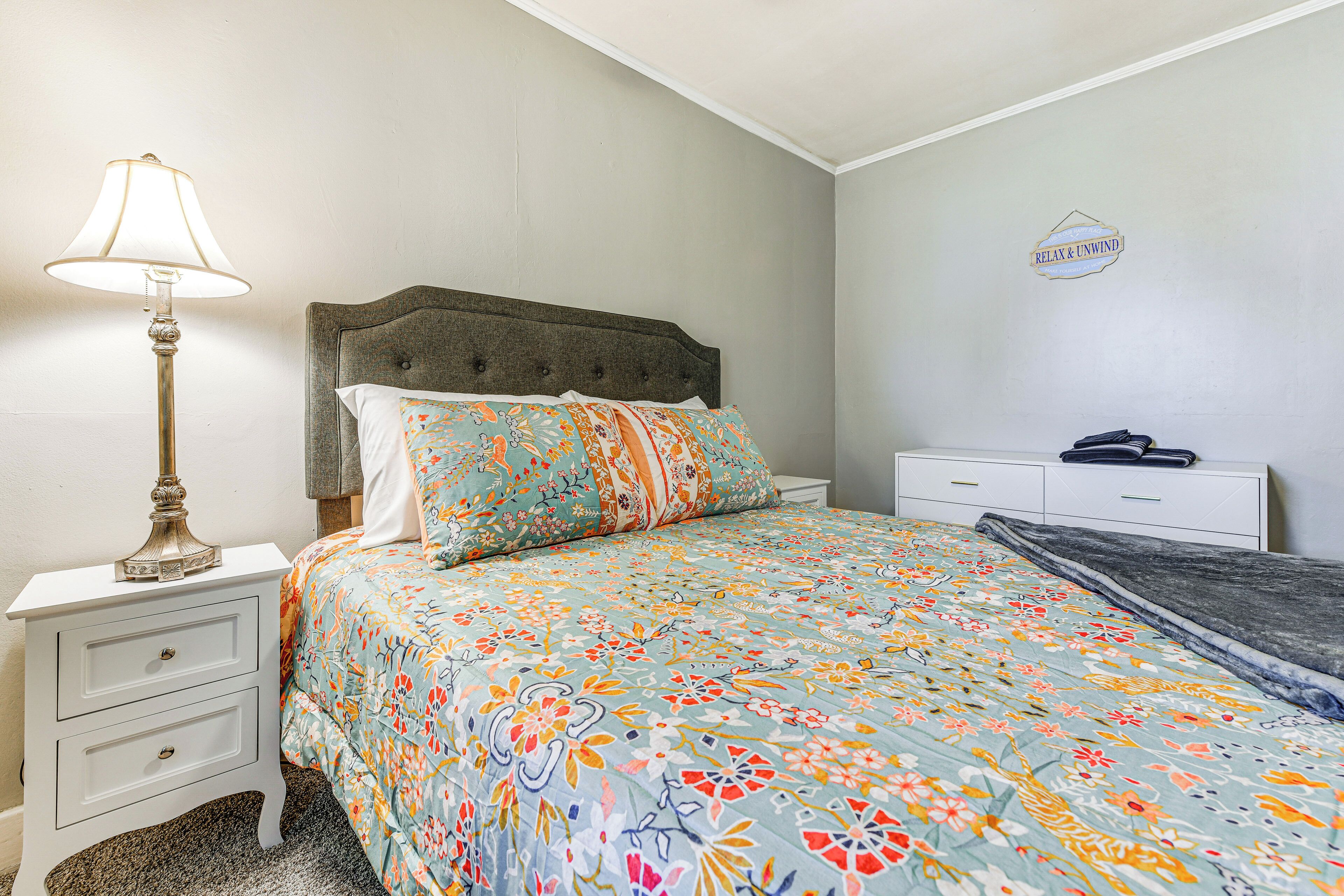A bedroom with a bed, a nightstand, a lamp, a dresser, and a wall sign.