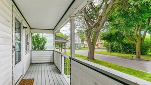 Un porche cubierto con piso de madera, barandillas blancas y vista a una calle con casas y árboles.