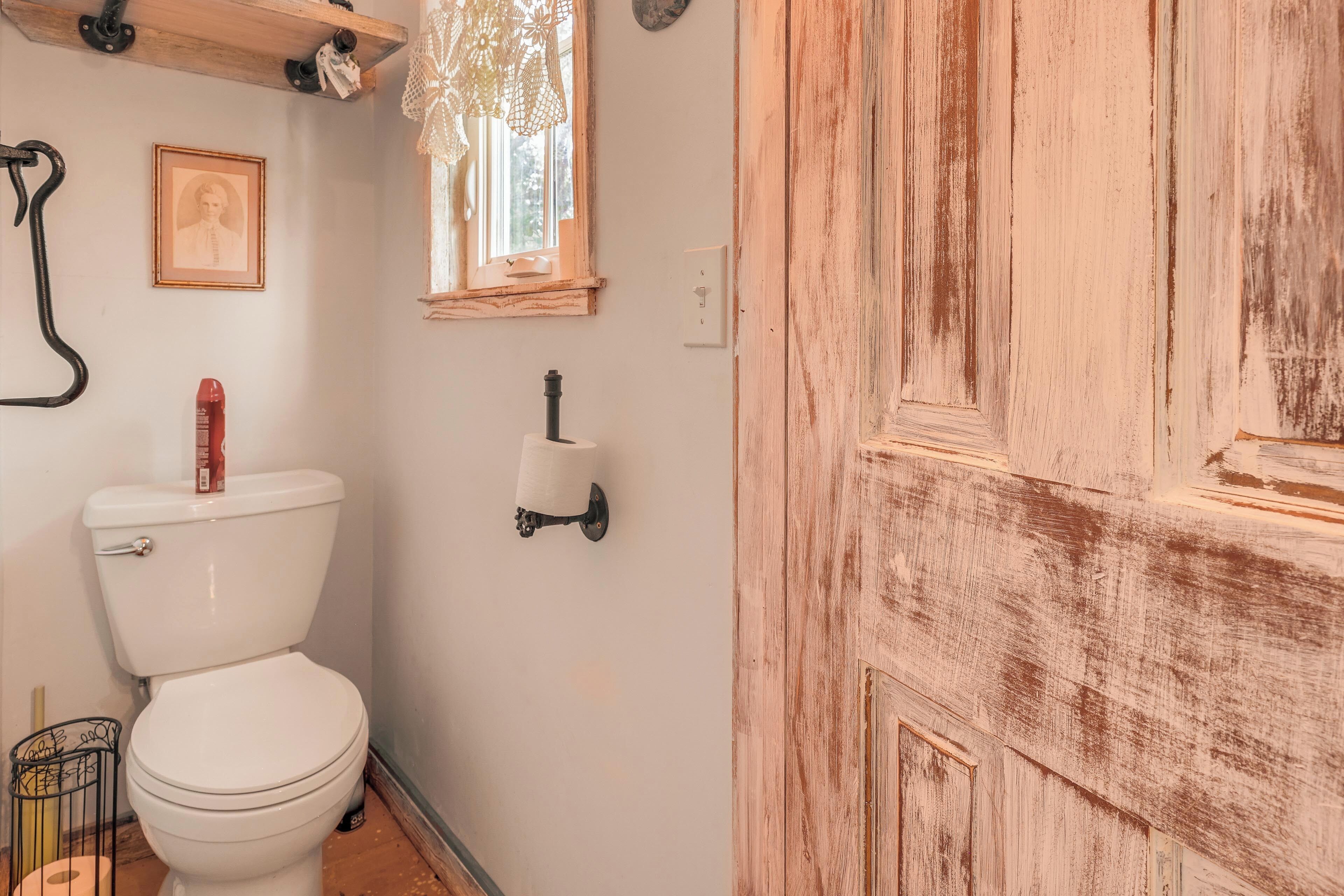 Une salle de bain avec des toilettes blanches, un rouleau de papier toilette, un tableau encadré accroché au mur et une porte en bois.
