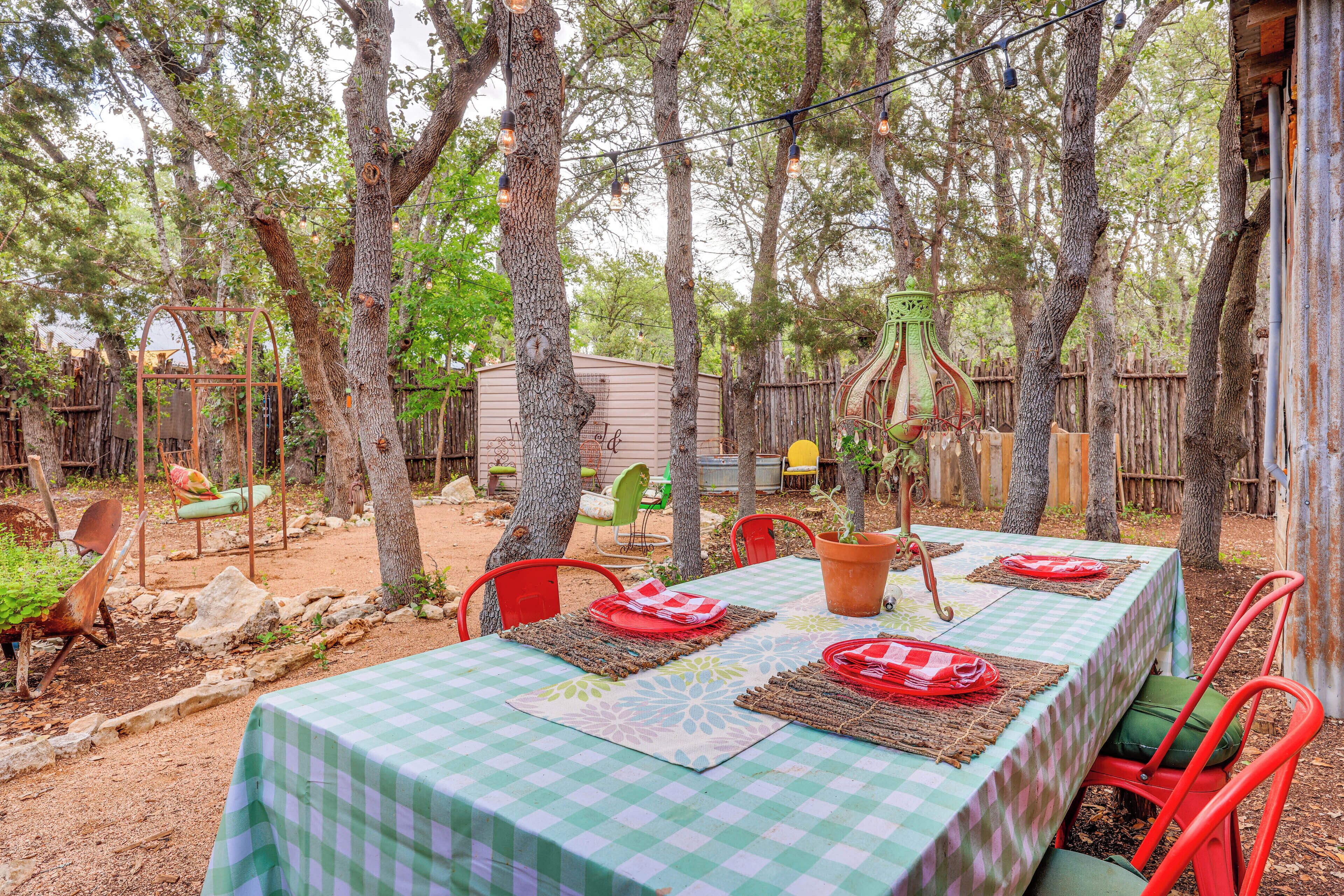 An outdoor dining area with a checkered tablecloth, red chairs, and a rustic setting with trees and a wooden fence.