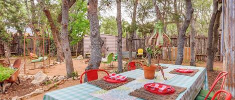 An outdoor dining area with a checkered tablecloth, red chairs, and a rustic setting with trees and a wooden fence.