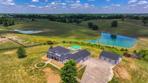 Une vue aérienne d’une grande maison dotée d’une piscine, entourée de vastes champs verdoyants et d’un petit lac.
