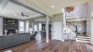 Un vaste salon doté d’une cheminée, d’un grand canapé d’angle gris et d’un plancher en bois franc. Un escalier mène à un niveau supérieur où se trouve un coin repas.