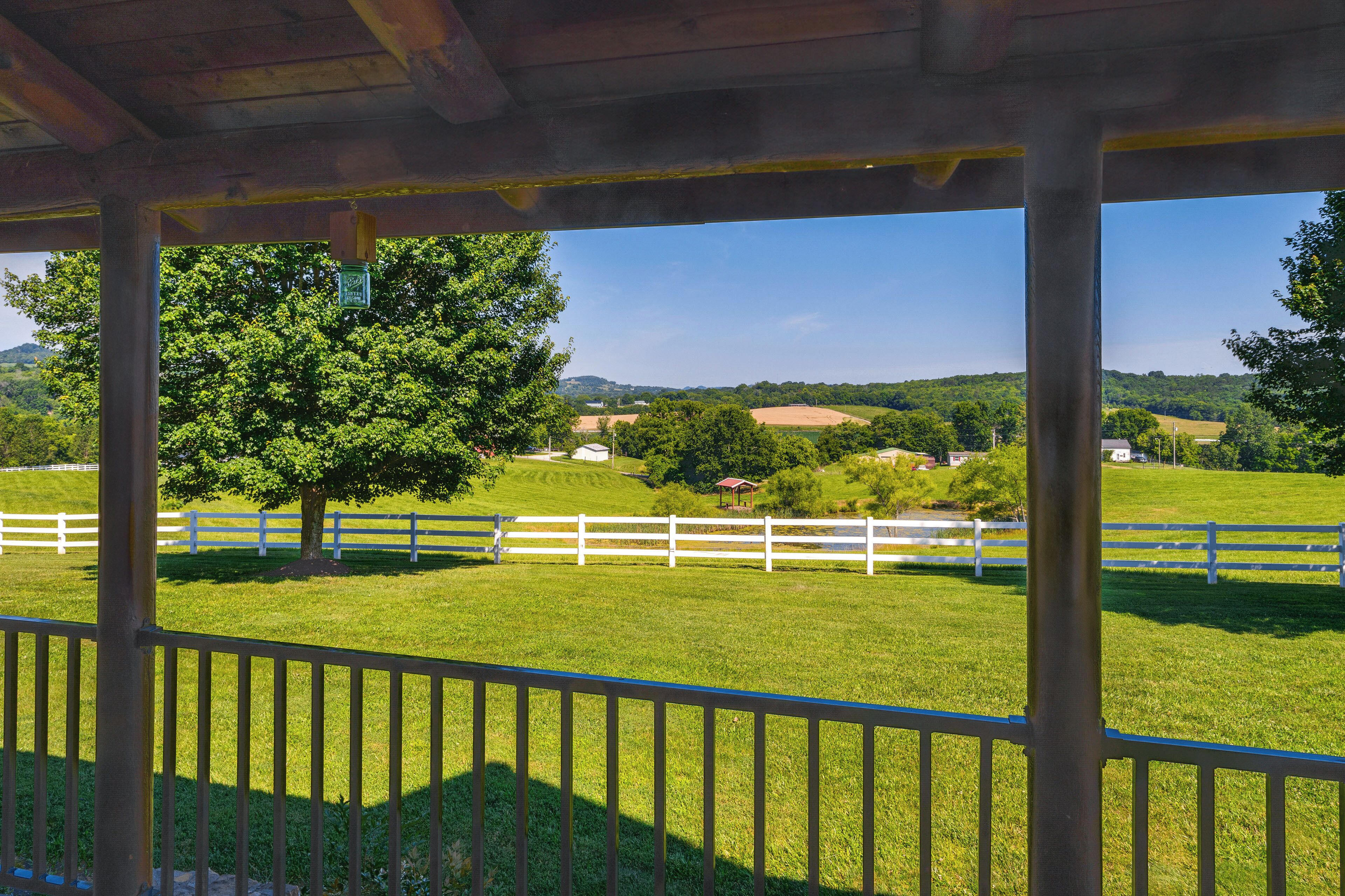 A covered outdoor area with a white fence, a large tree, and a view of a green field and distant buildings.