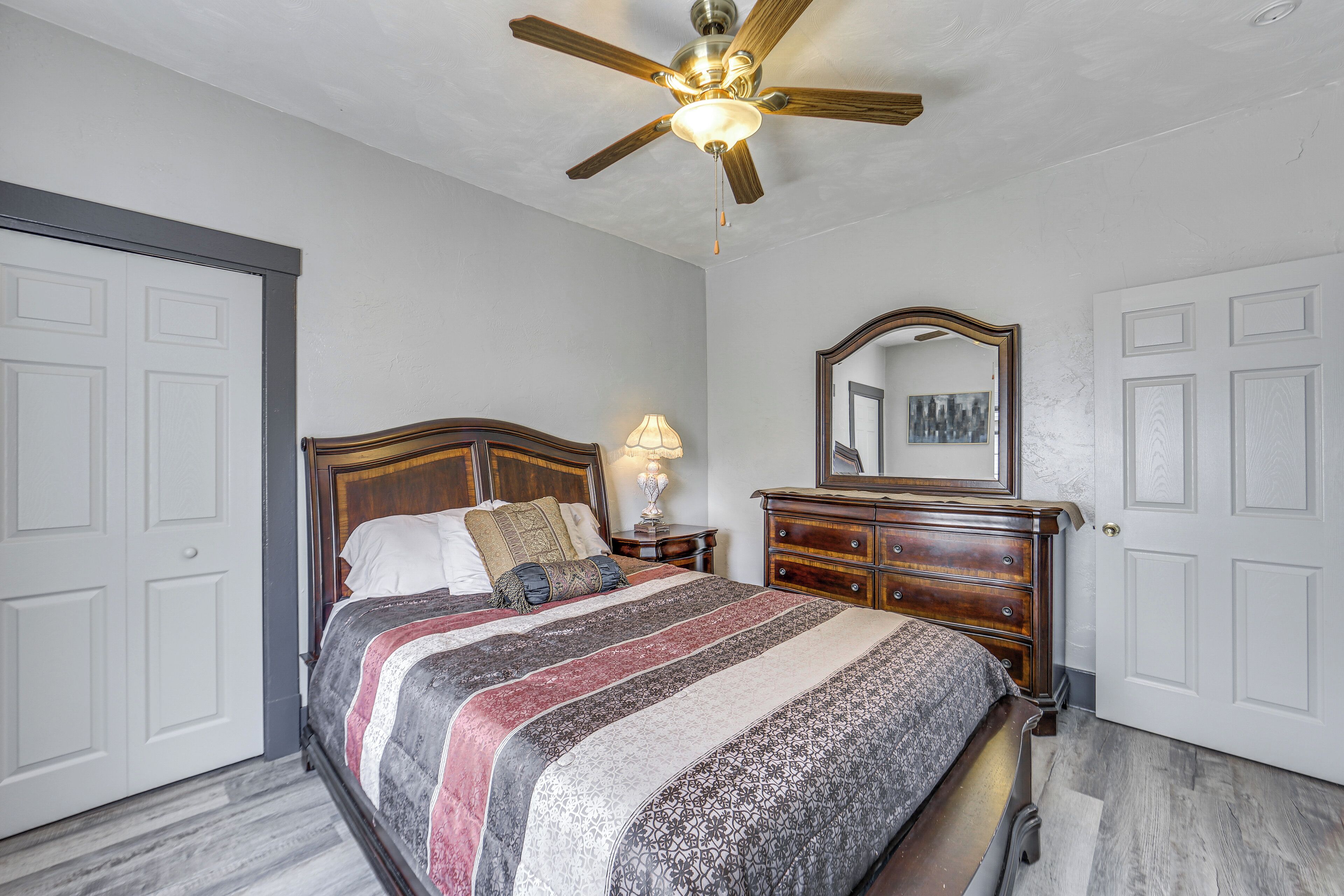 A bedroom with a wooden bed, a nightstand with a lamp, a ceiling fan, and a dresser with a mirror.