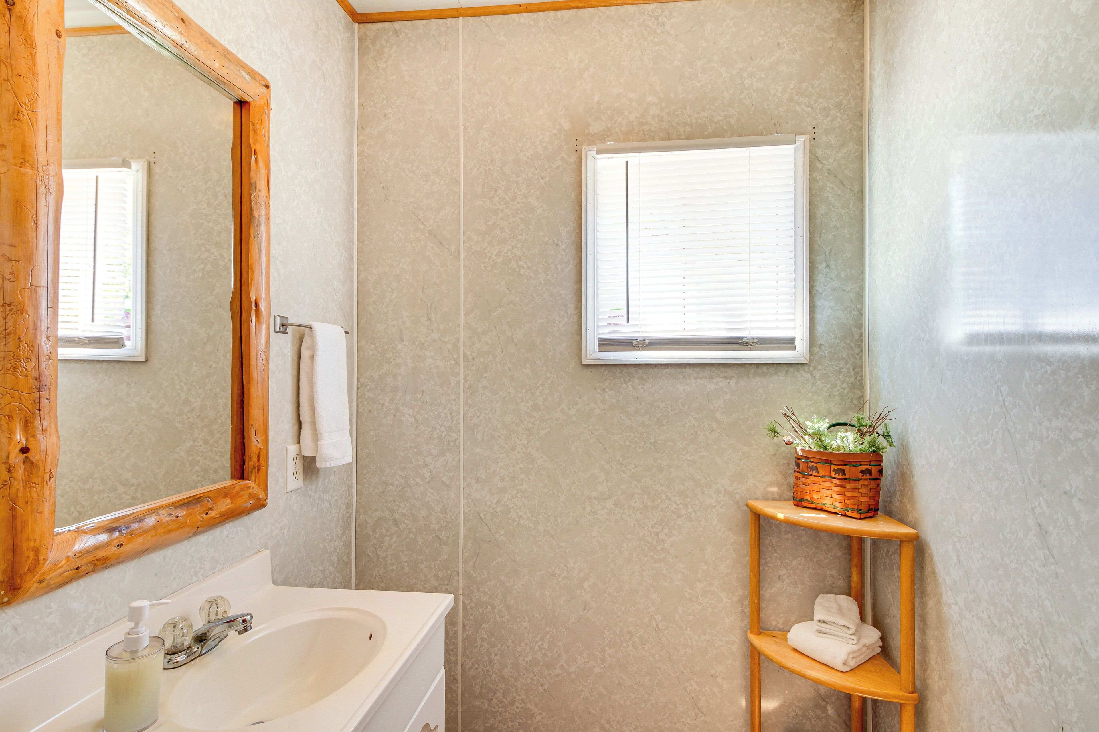 A bathroom with a wooden-framed mirror, a white sink with a soap dispenser, a towel rack, a window with blinds, and a wooden shelf with a basket.