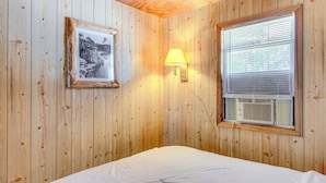 Una habitación con paneles de madera, una cama, un cuadro enmarcado, una lámpara de pared y un equipo de aire acondicionado.