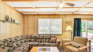Interior acogedor de una cabaña con techo de madera, un ventanal con persianas, un sofá, una mesa de madera y una silla.