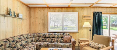 Interior acogedor de una cabaña con techo de madera, un ventanal con persianas, un sofá, una mesa de madera y una silla.