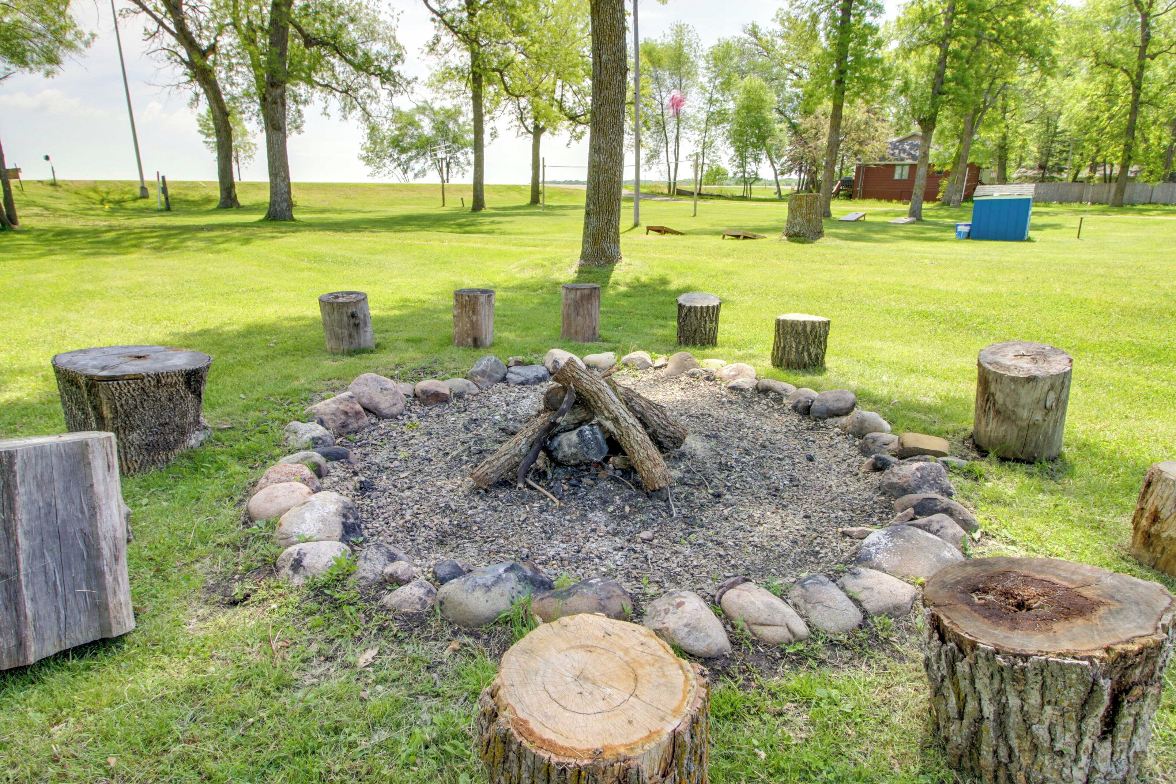 A fire pit surrounded by wooden stumps and rocks, set in a grassy area with trees and a building in the background.