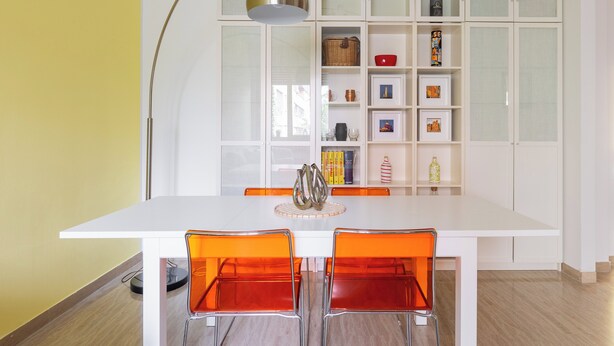 En matplats med ett vitt bord, orange stolar och ett skåp med glasdörrar som innehåller olika föremål.