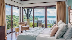 A bedroom with a large bed, two chairs, a small table, and a view of a lake and hills.