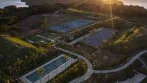 An aerial view of a resort with multiple tennis courts, a swimming pool, and a scenic ocean view.
