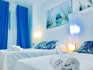A hotel room with two beds, blue curtains, a wall-mounted lamp, and a painting of a cityscape.