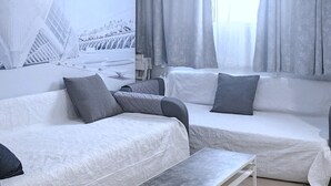 Ein modernes, minimalistisches Schlafzimmer mit zwei Betten, einem kleinen Tisch und einem großen Fenster mit Vorhängen.