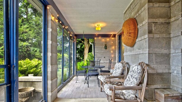 A covered outdoor seating area with wicker chairs, a stone wall, and a view of greenery.