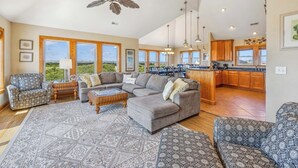 A spacious living room with a gray sectional sofa, patterned armchairs, and a large rug. A ceiling fan and pendant lights are visible. The kitchen area is visible in the background with wooden cabinets and a large window. - Soundfront Home w/ Indoor Pool, Hot Tub, Elevator, Game Room, Tiki Bar, Theater (Avon)