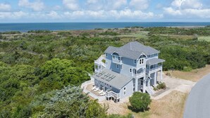 A large, two-story house with a balcony and a covered patio area, surrounded by dense greenery and overlooking a coastal landscape. - Soundfront Home w/ Indoor Pool, Hot Tub, Elevator, Game Room, Tiki Bar, Theater (Avon)