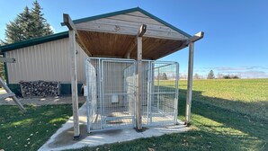 Una caseta metálica para perro con techo de madera y base de hormigón, situada en una zona con césped y un edificio al fondo.