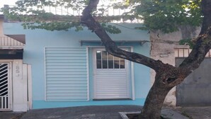 Un edificio de color azul claro, una puerta blanca con reja de metal y un árbol en primer plano.