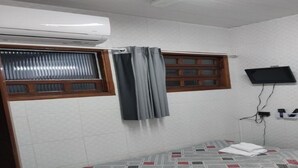 Una habitación pequeña y limpia con cama, ventana con cortinas, televisor montado en la pared y aire acondicionado.