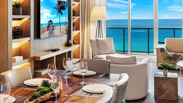 Ein Zimmer mit einem Holzesstisch, gedeckt für vier Personen, einem an der Wand befestigten Fernseher und einem großen Fenster mit Blick auf den Ozean.